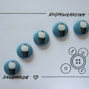 Könnte beinhalten: Vier blaue Stoffknöpfe mit einem weißen Kaktus-Design und dem Text "KNOPFWERKSTATT" und "HANDMADE" auf weißem Hintergrund.