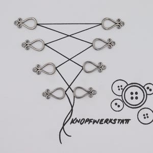 Puede incluir: Primer plano de seis lazos decorativos de metal plateado con un cordón negro enhebrado. Los lazos están dispuestos en dos filas de tres. El cordón está atado en un nudo en la parte inferior de la imagen. El texto "KNOFFWERKSTATT" está escrito en tinta negra debajo de los lazos.