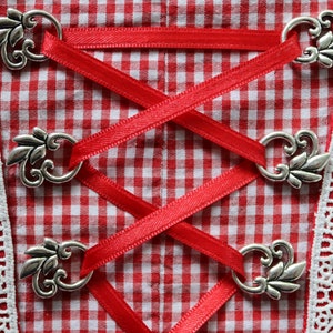 Puede incluir: Tela de cuadros rojos y blancos con un borde de encaje blanco. La tela está atada con una cinta roja y cierres de metal plateado.