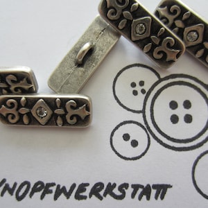 May include: Silver metal buttons with a decorative design and a clear rhinestone in the center. The buttons are rectangular and have a slightly raised design. The word "KNOPFWERKSTATT" is written in black on a white background.