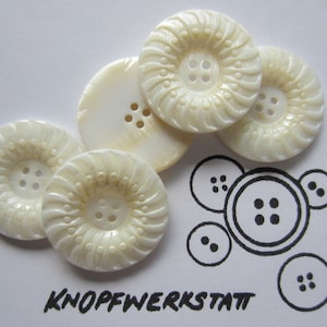 Può includere: Cinque bottoni decorativi rotondi e bianchi con un motivo a spirale e quattro fori ciascuno. I bottoni sono disposti a semicerchio su una superficie bianca. La parola "KNOFFWERKSTATT" è scritta in inchiostro nero sotto i bottoni.