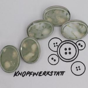 Könnte beinhalten: Fünf ovale Knöpfe mit einem grün-weißen Marmormuster. Die Knöpfe haben einen weißen Rand und sind halbkreisförmig angeordnet. Der Text "KNOFFWERKSTATT" ist in schwarzer Tinte unter den Knöpfen geschrieben.
