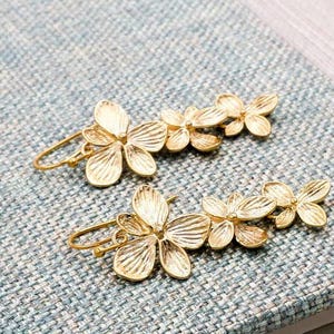 May include: A pair of gold-colored floral earrings. Each earring features a cluster of delicate, stylized flowers. The earrings are set against a textured, light blue surface, showcasing their intricate design.