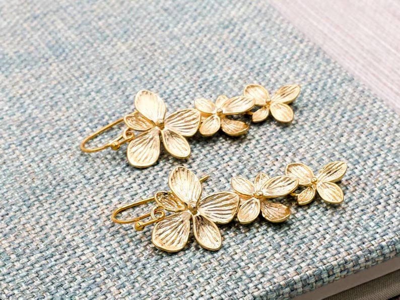 May include: A pair of gold-colored floral earrings. Each earring features a cluster of delicate, stylized flowers. The earrings are set against a textured, light blue surface, showcasing their intricate design.