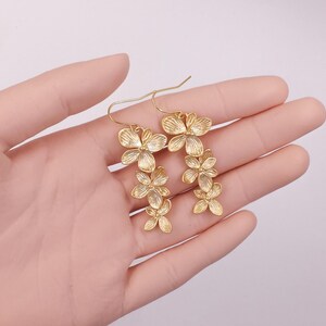 May include: Gold-colored floral earrings. Each earring features a cascade of delicate, flower-shaped elements. The earrings are designed with a hook closure. The earrings are displayed against a soft, neutral background.