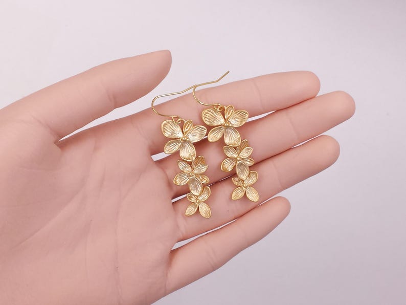 May include: Gold-colored floral earrings. Each earring features a cascade of delicate, flower-shaped elements. The earrings are designed with a hook closure. The earrings are displayed against a soft, neutral background.