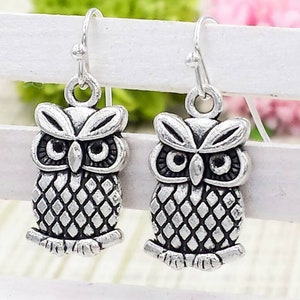 May include: A pair of silver owl dangle earrings with a detailed design. The owls have a textured pattern and black eyes.