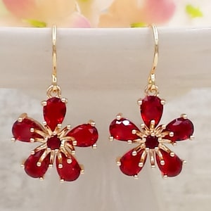 May include: A pair of gold dangle earrings with red flower-shaped charms. Each charm has five red pear-shaped stones and a smaller red round stone in the centre.