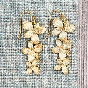 May include: A pair of gold-colored floral earrings. Each earring features a cascade of three stylized flower clusters, suspended from a simple hook. The earrings are set against a textured, light blue background.