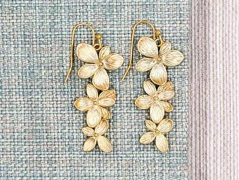 May include: A pair of gold-colored floral earrings. Each earring features a cascade of three stylized flower clusters, suspended from a simple hook. The earrings are set against a textured, light blue background.