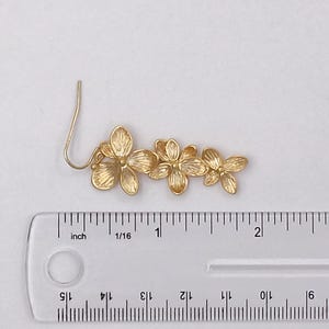 May include: A gold-colored floral earring with a hook, featuring three flower blossoms. The earring measures approximately 2.5 inches in length. The flowers have detailed petal designs, and the earring is set against a white background.