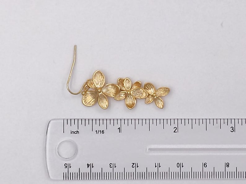 May include: A gold-colored floral earring with a hook, featuring three flower blossoms. The earring measures approximately 2.5 inches in length. The flowers have detailed petal designs, and the earring is set against a white background.
