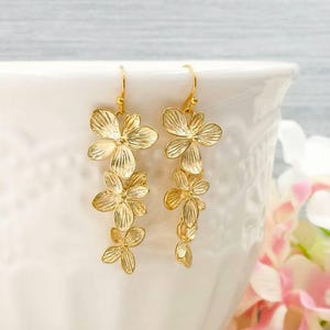 May include: Gold-colored floral drop earrings. Each earring features multiple flower-shaped pendants, creating a cascading effect. The earrings are suspended from a simple hook. The background is a soft white and pink, suggesting a delicate and feminine aesthetic.