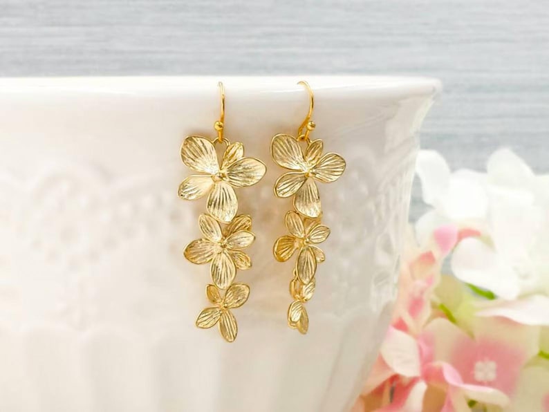 May include: Gold-colored floral drop earrings. Each earring features multiple flower-shaped pendants, creating a cascading effect. The earrings are suspended from a simple hook. The background is a soft white and pink, suggesting a delicate and feminine aesthetic.