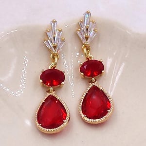 May include: Gold-toned drop earrings featuring a cluster of clear crystals at the top, a small oval red gemstone, and a larger teardrop-shaped red gemstone. The earrings are set against a light-colored background.