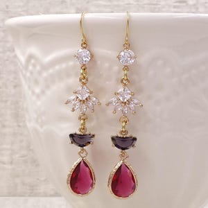 May include: A pair of gold-tone drop earrings featuring a clear round crystal at the top, a fan-shaped cluster of clear marquise crystals in the center, a dark purple semi-circular stone, and a vibrant pink teardrop-shaped crystal at the base.