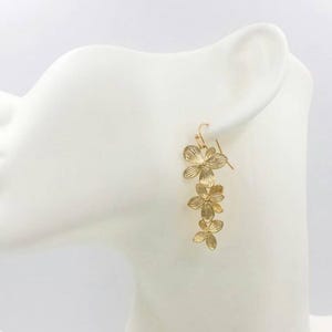 May include: Gold-colored floral drop earrings. The earrings feature a cascading design of three flower clusters, each with detailed petal patterns. The earrings are displayed on a white mannequin head.