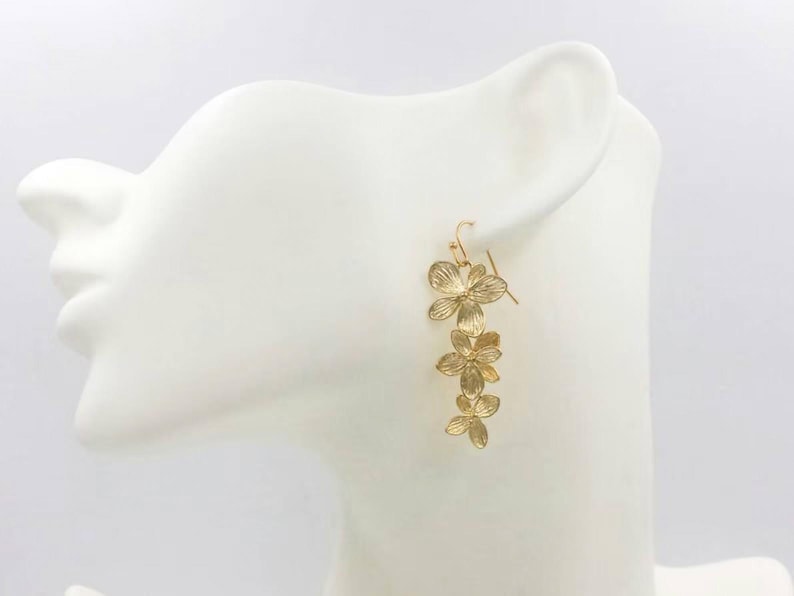 May include: Gold-colored floral drop earrings. The earrings feature a cascading design of three flower clusters, each with detailed petal patterns. The earrings are displayed on a white mannequin head.