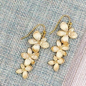 May include: A pair of gold-colored floral drop earrings. Each earring features a cascade of three flower clusters, with delicate petals and a hook closure. The earrings are displayed on a textured blue and white surface.