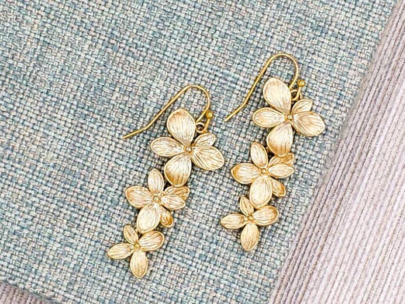 May include: A pair of gold-colored floral drop earrings. Each earring features a cascade of three flower clusters, with delicate petals and a hook closure. The earrings are displayed on a textured blue and white surface.
