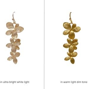 May include: Gold-colored floral earrings. Each earring features three flower-shaped pendants hanging from a hook. The image shows the earrings in two different lighting conditions: ultra bright white light and warm light dim tone.
