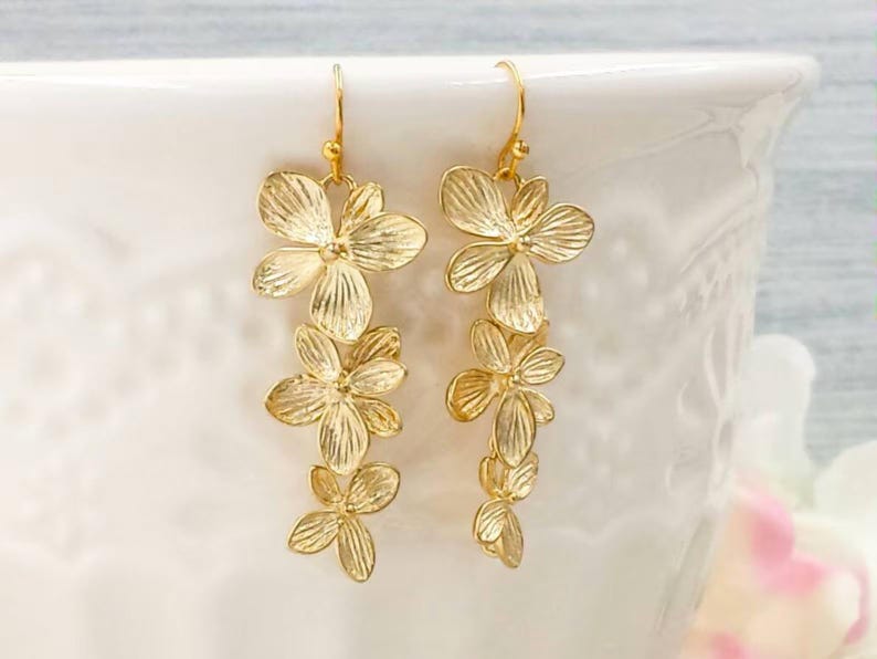 May include: A pair of gold-colored floral drop earrings. Each earring features three cascading flower clusters with detailed petal designs. The earrings have a hook closure and are displayed against a white background.