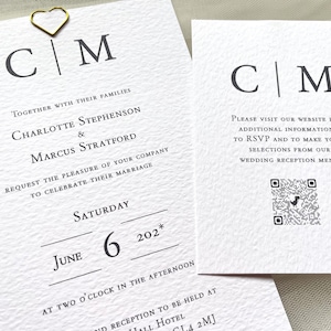 May include: A set of wedding stationery on textured white paper. The design features the initials "CM" in a serif font, along with the names Charlotte Stephenson and Marcus Stratford. A gold heart-shaped paper clip accents the top. Includes a QR code.