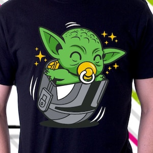 Puede incluir: Una camiseta negra con una imagen de dibujos animados de un bebé Yoda verde que lleva un chupete y está sentado en una cápsula.
