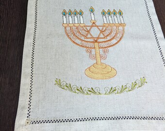 Può includere: Un runner da tavolo in lino bianco con una menorah e una stella di David ricamate. La menorah è dorata con nove candele, e la stella di David è al centro della menorah.