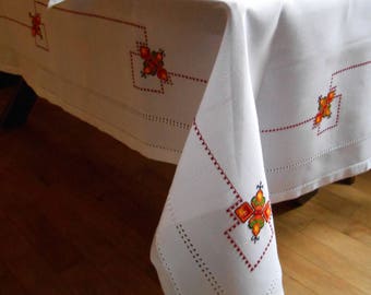 Può includere: Tovaglia bianca con ricami floreali rossi e arancioni. La tovaglia è stesa su un tavolo di legno con piatti bianchi sopra.