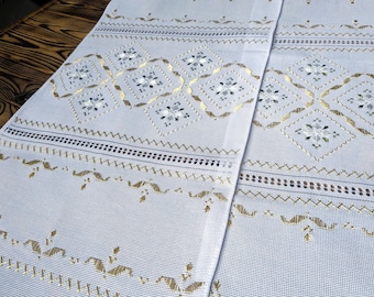 Può includere: Un tessuto azzurro con intricati motivi ricamati in oro e bianco. Il design presenta forme geometriche, motivi floreali e bordi decorativi. Il tessuto è piegato, mostrando la lavorazione e la trama dettagliate.