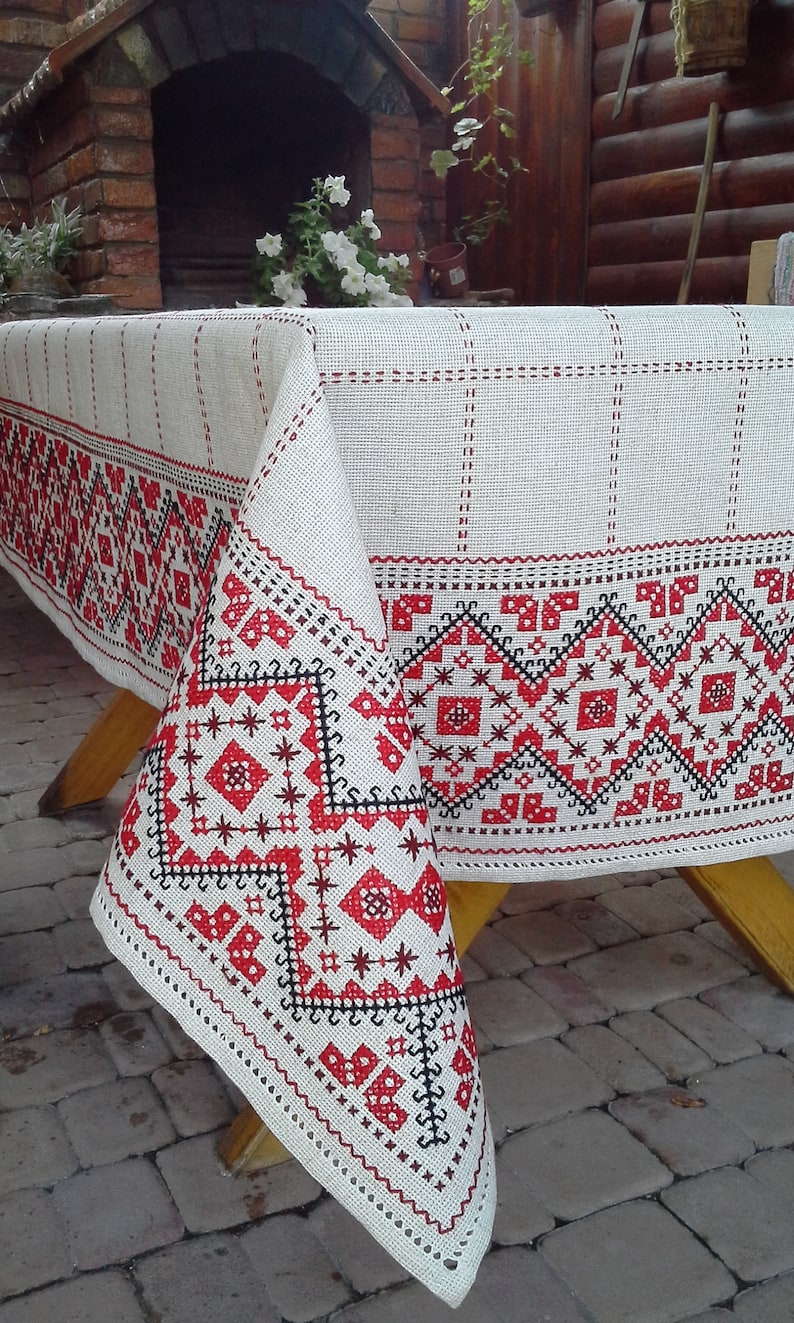Pu&ograve; includere: Un tovaglia di lino bianco con un bordo ricamato rosso e nero. Il ricamo presenta motivi geometrici e stelle.