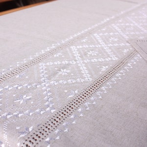 May include: A white linen tablecloth with a delicate embroidered pattern of white flowers and geometric shapes. The tablecloth has a subtle sheen and a soft texture.