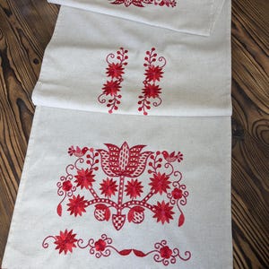 May include: A white linen table runner with red embroidered floral designs. The runner has a fringe on the bottom edge.