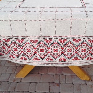 Pu&ograve; includere: Un tovaglia di lino bianco con un bordo ricamato rosso e nero. Il bordo presenta un motivo geometrico con quadrati e triangoli ripetuti.