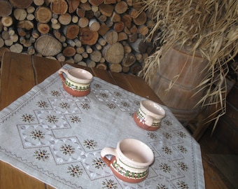 Può includere: Tre tazze in ceramica marrone e bianca con disegni verdi e marroni sono su una tovaglia bianca con un motivo ricamato. La tovaglia è su un tavolo di legno.