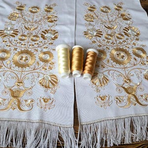 May include: White fabric panels with gold floral embroidery and fringe trim. Three spools of gold thread are placed on the fabric. The embroidery features stylized flowers and leaves.