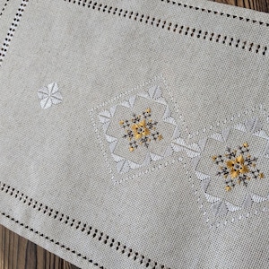 Pode incluir: Um corredor de mesa de linho bege com um design bordado branco e dourado. O design apresenta um padrão repetitivo de formas geométricas e flores.