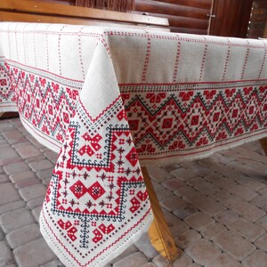 Pu&ograve; includere: Una tovaglia di lino bianco con motivi geometrici ricamati in rosso e nero. La tovaglia &egrave; drappeggiata su un tavolo di legno e ha un bordo a frangia.