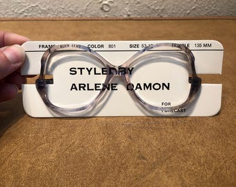 May include: A white card with the text "Styled by Arlene Damon for Forecast" and a pair of clear plastic glasses with a gray tint. The card also includes the text "Frame: A147100, Color: 801, Size: 53-15, Temple: 135 MM."