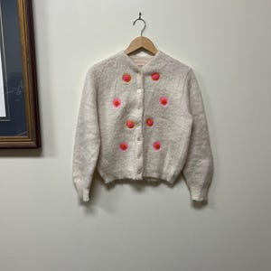 May include: A cream-colored, button-down cardigan sweater with bright pink and orange floral embroidery. The sweater is hanging on a wooden hanger against a white wall. The embroidery is arranged in a symmetrical pattern.