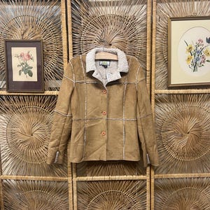 May include: A tan suede jacket with a cream-colored shearling collar and trim. The jacket has three brown buttons down the front and is hanging on a wooden hanger. The background features a woven wall decor and framed artwork.