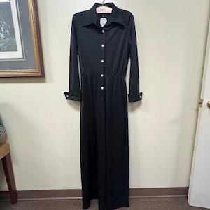 Könnte beinhalten: Ein langer, schwarzer Jumpsuit mit Kragen und langen Ärmeln. Der Jumpsuit hat weiße Knöpfe auf der Vorderseite und an den Manschetten. Das Kleidungsstück hängt an einem Holzbügel.