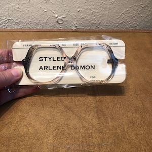 May include: A pair of clear, brown-tinted eyeglasses with a rounded, rectangular frame. The glasses are packaged in a plastic bag with a white card that reads "Styled by Arlene Damon" and includes the frame size and measurements.