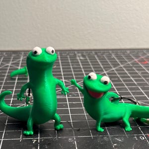May include: Two green lizard figurines with white eyes and black pupils. One lizard is standing upright, while the other is in a playful pose with its mouth open. Both have small black cords attached, suggesting they are keychains or charms.
