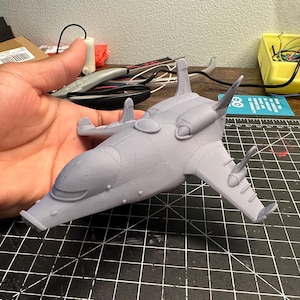 Può includere: Modello grigio chiaro stampato in 3D di un velivolo futuristico. Il modello presenta ali, cabina di pilotaggio e dettagli aerodinamici. È tenuto in mano su uno sfondo di strumenti e un tappetino da taglio.