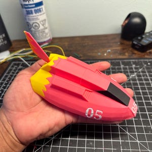 Puede incluir: Un modelo de nave espacial impreso en 3D de color rosa y amarillo con un cono de nariz negro y el número "05" impreso en el lateral.