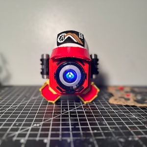 May include: A red and white robot with a blue light in its eye. The robot is wearing a white hat with a black and brown patch on it. The robot is standing on a black and gray grid.