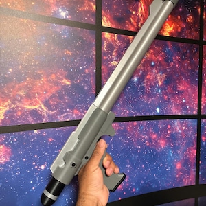 May include: A silver and gray prop gun with a black tip and gray handle. The prop gun is held in a person's hand. The background is a colorful galaxy scene.