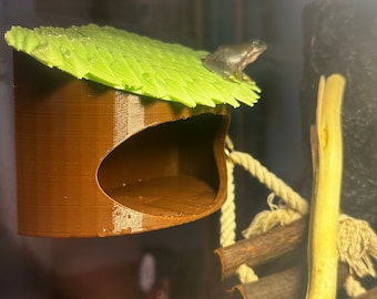 May include: A brown, 3D printed frog house with a green leaf roof. The house is hanging from a rope and a small frog is sitting on the roof.